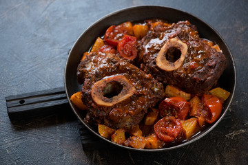Metal pan with osso buco stewed with oranges and tomatoes, dark brown stone background, studio shot
