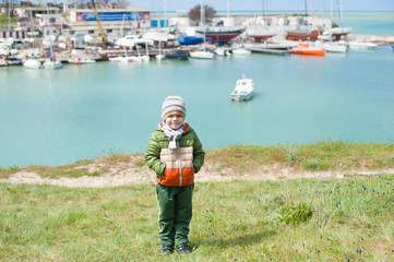 happy little boy in warm jacket and hat and scarf on green slope against background of sea bay with ships in early spring