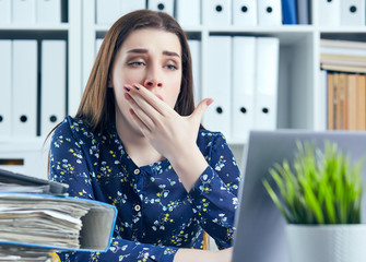 Young business woman yawning at a modern office desk in front of laptop, covering her mouth....