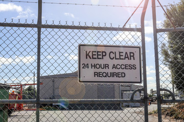 a black and white Keep Clear, 24 Hour Access Required sign on the padlocked gates of a work site, with lens flare