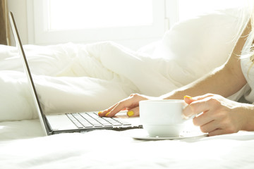 Fototapeta na wymiar Close up, young blond woman in bed, white sheets linens holding cup of morning coffee, surfing browsing blogging eshopping on laptop. Perks of freelance concept. Blond female in tight shirt Background