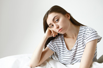 Fototapeta premium Pretty girl in striped pajamas having bored unhappy expression, looking at camera with hand under her cheek while sitting on bed at white wall with copy space for your text or advertising content