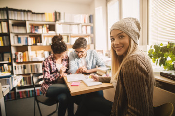 Fototapeta premium Photo of a gorgeous blond teenage student girl sitting in the library with two friends and doing homework.