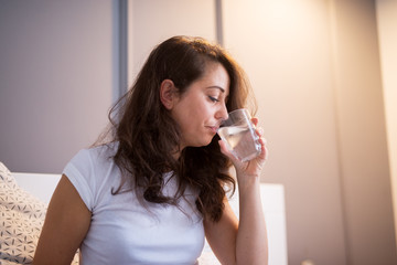 Beautiful middle aged woman drinking water while sitting on the bed in pajamas.