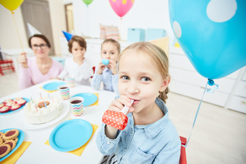 Happy cute girl with whistle looking at camera while sitting by festive table on background of her...