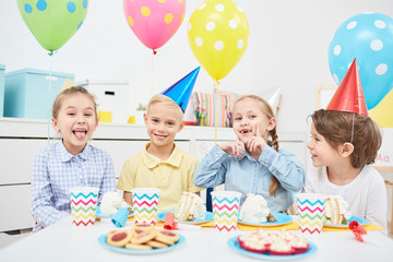 Funny little kids in birthday caps enjoying party with balloons and tasty desserts in kindergarten