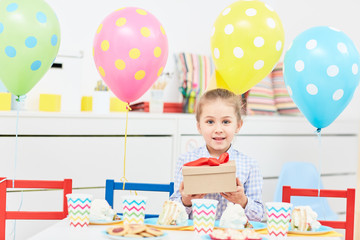Fototapeta premium Pretty little girl showing box with red ribbon containing her birthday gift while sitting by festive table
