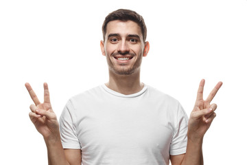 Indoor portrait of young male isolated on white background in blank tshirt with optimistic smile, showing victory sign with both hands, looking friendly and willing to welcome and communicate