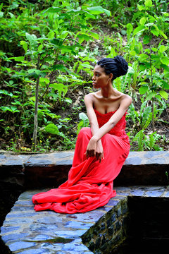 African-American woman sitting on a background of green plants in the rocks in a red dress