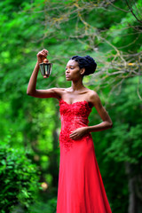 Obraz na płótnie Canvas African American girl in a red dress, with dreadlocks, with a lamp and a candle in his hand, stands in the Park in the summer on a background of green plants and looks at the fire. Vertical view