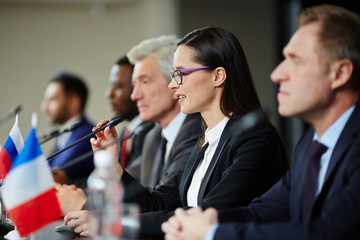 Young female politician in formalwear talking in microphone while making report at political...