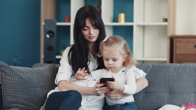 Happy mother hug daughter use phone together sit on sofa woman baby hand love child person girl kid internet cartoon technology smartphone face touchscreen mobile looking application touch game play