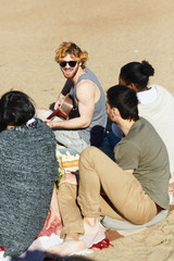 Group of friends listening to guy playing guitar and singing during picnic on sand