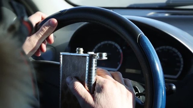 Alcohol is driving a car while driving. The hand of a drunk driver holds a flask with an alcogol at the helm of his car.