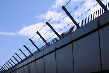 Safety wall with barbed wire and spikes on top of a fence provide security - blue sky background...
