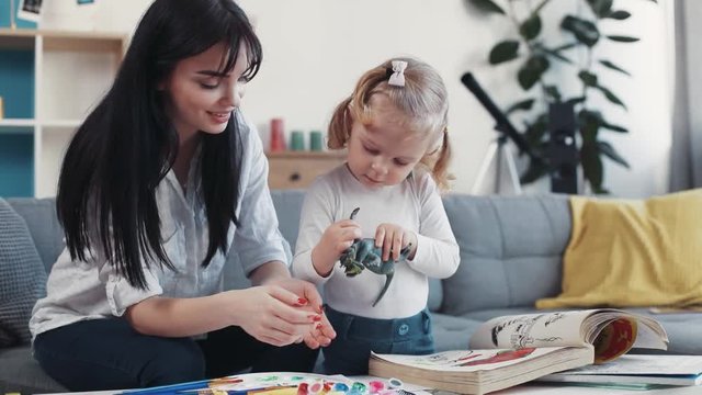 little child painting on paper book with mum hug sit on sofa together at home happy kid family hobby girl love house coloring mom play art childhood table artist closeup mother