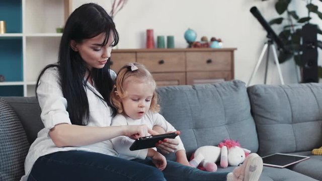 Mother with daughter sit on sofa use phone look some interesting hug woman baby hand love child girl kid internet cartoon technology smartphone face home mobile touch mom game little play application