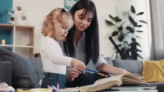mum with little child paitning on papper book hug sit on sofa together at home happy kid family hobby girl love house coloring mom play art childhood table artist closeup mother