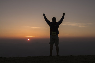 Man with backpack putting his hands up  and standing on cliff at sunset time