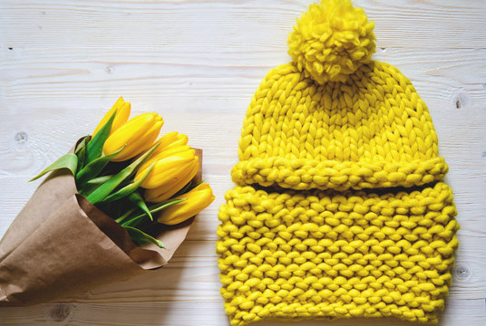 A yellow knitted hat and a snore from thick Merino yarn. Nearby is a bouquet of yellow tulips wrapped in paper.
