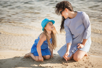 Mother and her little daughter having fun at the coast. Young pretty mom and her child playing near the water and drawing heart on the sand