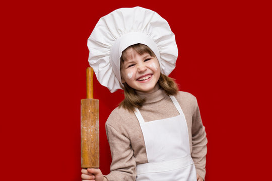 Portrait of happy little girl in chef uniform holds a rolling pin isolated on red. Kid chef. Cooking Process Concept 