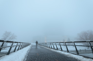 Fuzzy silhouette of a man passing through a pedestrian bridge and reflected in puddles obscure in dense fog . Mystic background