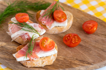 Italian prosciutto and feta cheese bruschetta decorated by tomato and dill. Antipasti on grilled baguette bread.