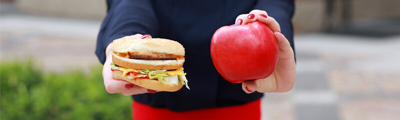 What to choose. healthy and unhealthy food, female hands holding a hamburger and an apple, standing on the street, close-up.
