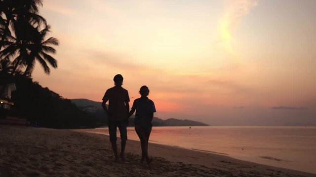 Silhouette of young couple walks on the beach listen to music with headphones at amazing sunset holding hands. Happy young couple enjoying ocean sunset during travel vacation. slow motion. 1920x1080