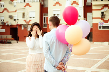 Happy couple outdoors man prepared surprise for a woman. Woman eyes closed, man hiding balloons at his back.
