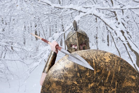 Spartan attacks someone with sharp metallic spear in winter forest. He wers traditional spartan armor: spear, shield, helmet, red long cape. Man is careful and bold. Warrior has big blue eyes.