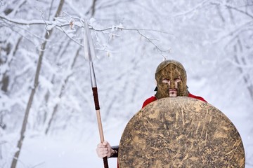 Spartan keeping spear and shield in snowy forest. He is wearing red long cape is stealing through...
