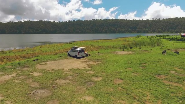 Aerial view circle around white camper van near lake, two people sitting at table under canopy