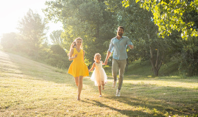 Naklejka na ściany i meble Full length of two happy parents with a healthy lifestyle, wearing casual clothes while running together with their cute daughter in a sunny day of summer