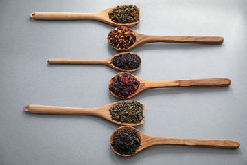 Spoons with different types of dry tea leaves on grey background