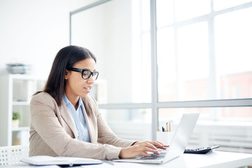 Secretary in eyeglasses typing on laptop at office