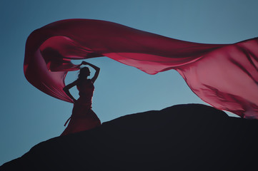 young woman in a red dress with light fabric flying in the wind at rock on background of blue sky 01