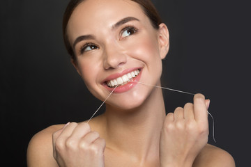 Portrait of excited cute girl is standing and using dental floss for cleaning teeth. She is looking aside with joy and smiling. Isolated on dark background. Dental hygiene concept