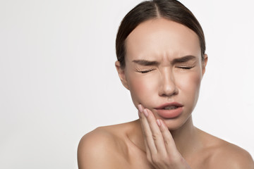 Fototapeta premium Dental pain. Portrait of joyless young woman is standing and holding her hand on cheek. She is closed eyes while wincing her face in terrible pain. Isolated background and copy space in the left side
