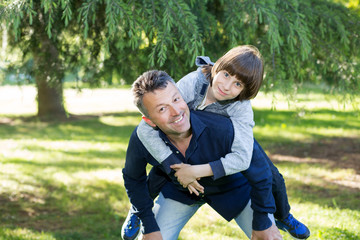 Fototapeta premium Portrait of father with his son having fun in summer park. Piggyback. Family fun. Happy boy playing with dad summer nature outdoor. Parenting. Two generations.