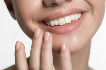 Close up of smiling mouth and fingers of happy young lady enjoying her fresh skin. She is slightly touching her chin and smiling with pleasure. Isolated