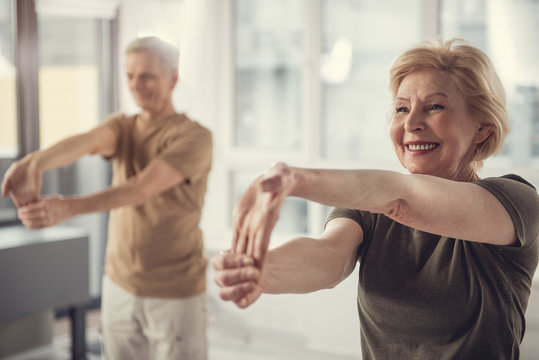 Waist up portrait of content mature female doing hand stretching exercise. Elderly man standing beside her in same posture. Focus on lady