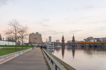 Fototapeta na wymiar Scenery and pedestrian way of riverside along Spree river and East side gallery with Oberbaumbrücke behind during sunset in Berlin, Germany.