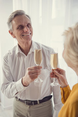 Enjoyed retired man and woman standing in living room and going clinking glasses with champagne