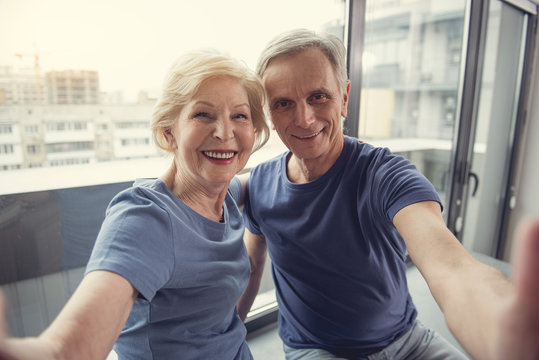 Waist up portrait of contented mature man and woman hugging and taking photo of themselves on windowsill