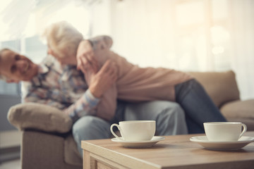Focus on mugs on desk. Laughing old man and woman lying on sofa and hugging