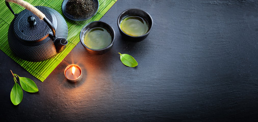 Grüner Tee in asiatischer Teekanne aus Eisen mit Blättern und Bambusmatte auf schwarzem Stein
