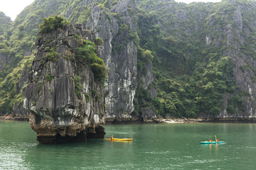 Enormous towering limestone islands over emerald water with tourists kayaking under the void of island in summer at Quang Ninh, Vietnam.
