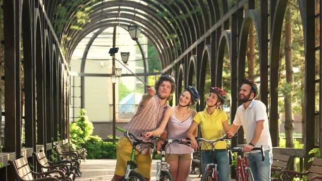 Four beautiful friends with monopod outdoors. Young cheerful male and female cyclists taking a photo with selfie stick.
